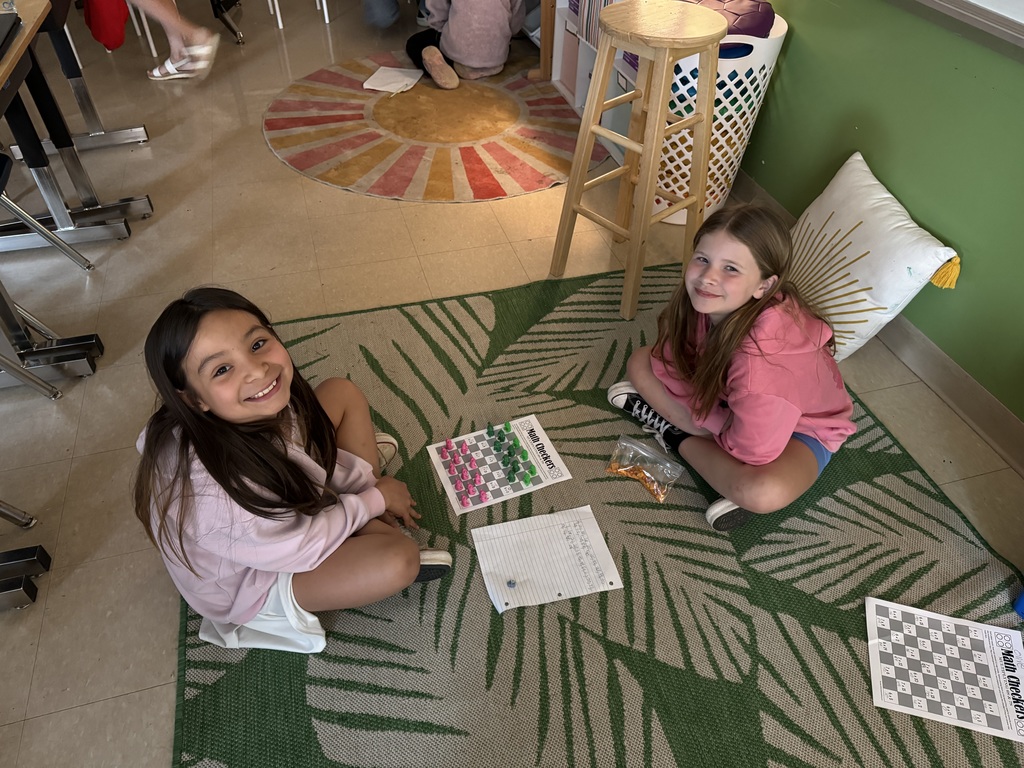 Two girls sit on a green rug, smiling while playing a math board game with colorful pieces and a worksheet nearby.