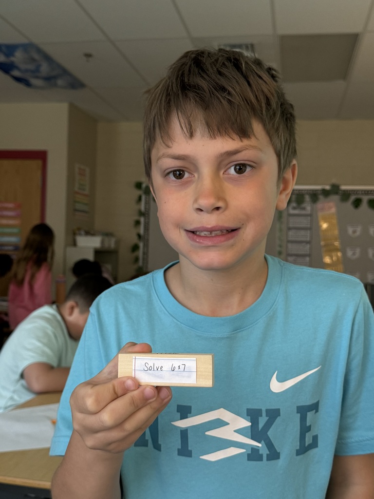 A boy holds up a small block labeled “Solve 6 × 7,” smiling slightly in a classroom setting.