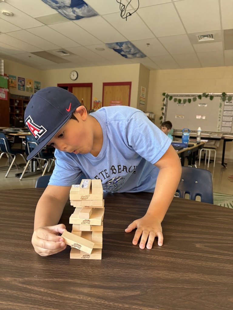 A boy wearing a cap carefully pulls a block from a Jenga tower at a classroom table.