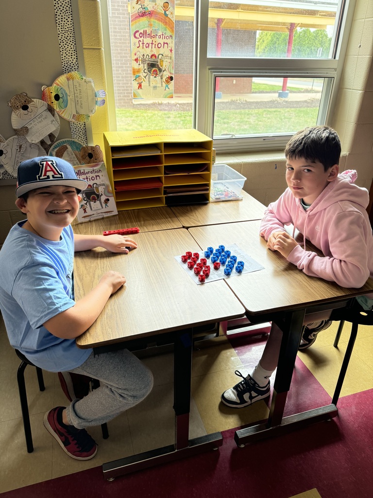 Two boys sit at a table by a window, playing a math board game with red and blue pieces; one smiles while the other looks focused.