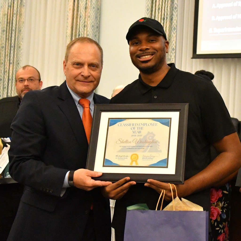 A man in a suit presents a framed certificate to another man wearing a black polo shirt and cap. Both are smiling and holding the award together, which reads “Classified Employee of the Year 2026–2027” and is awarded to Shelton Washington. The recipient also holds a gift bag