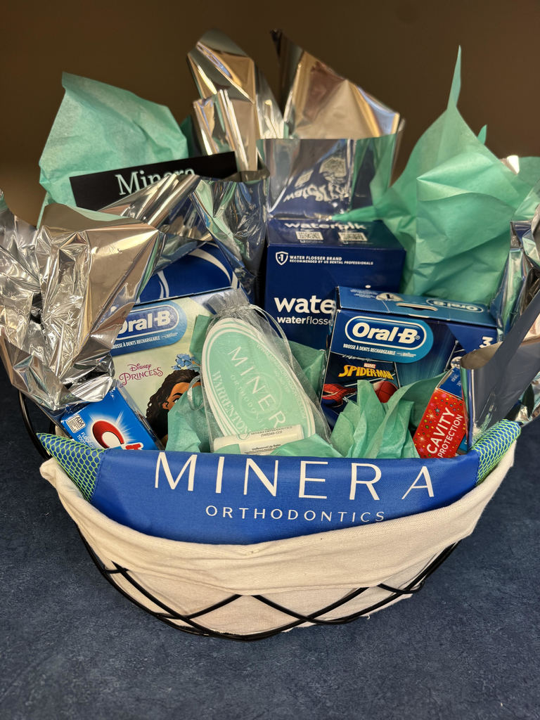 A gift basket filled with dental‑care items from Minera Orthodontics. The basket includes Oral‑B toothbrushes featuring Disney Princess and Spider‑Man designs, a box labeled “water flosser,” a circular container with the Minera Orthodontics logo, and a folded branded item in teal and silver tissue paper. The items are arranged neatly inside metallic wrapping.