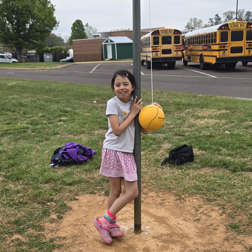 Fourth grader Sarah H proudly standing next to a newly hung tetherball with school buses in the background