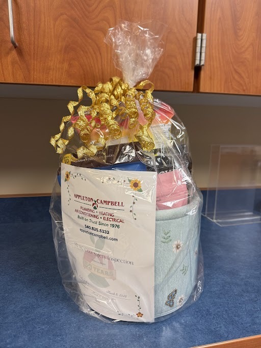 A gift basket wrapped in clear plastic with a gold ribbon on top. Inside the basket are several different snack bags of sweet and salty treats. Attached to the front is a printed card displaying the Appleton Campbell logo and services—plumbing, heating, air conditioning, and electrical—along with the slogan “Built on Trust Since 1976,” a phone number, website, and a note about a complimentary inspection. The date “March 8, 2018” appears at the bottom of the card.