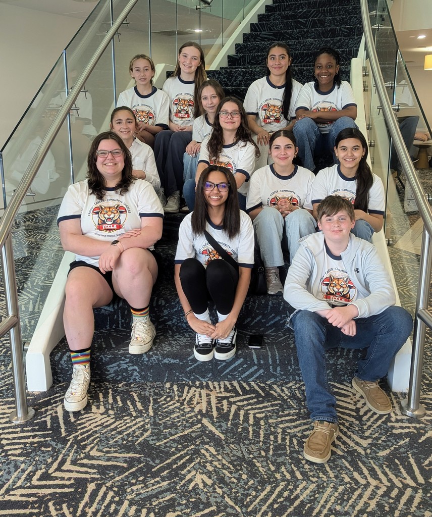 A group of middle school students wearing matching FCCLA T-shirts sit together on a carpeted staircase inside a modern building with glass railings. They are arranged in several rows, smiling at the camera, with a mix of casual outfits like jeans, sneakers, and hoodies. The setting appears bright and welcoming, suggesting a conference or event venue.