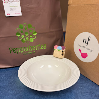 A white ceramic bowl with a small decorative attachment shaped like a colorful beach bag clipped to the rim. Behind the bowl are two shopping bags: one brown with a white circular “nora fleming” logo featuring a pink bird silhouette, and one kraft bag with a green “Pepperberries Palmyra, PA” logo and an attached business card. The display appears to showcase a Nora Fleming serving piece and its miniature accessory.