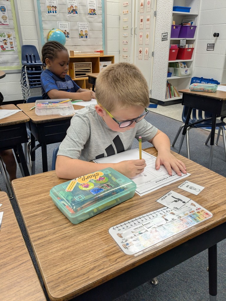 A student wearing glasses focuses on writing with a pencil at their desk while another student works behind them in a classroom.