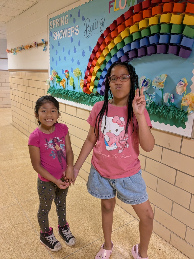 Two students stand in a hallway holding hands and smiling in front of a colorful bulletin board with a rainbow and spring theme.