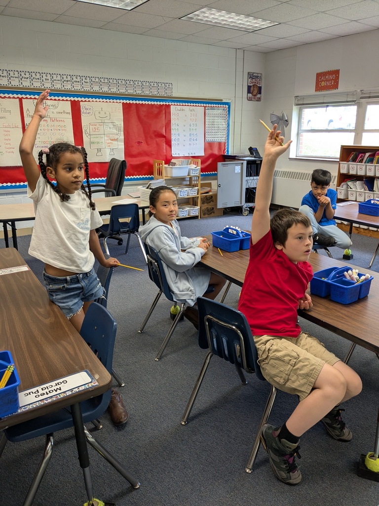 Students sit at desks in a classroom, raising their hands to answer a question during a lesson.