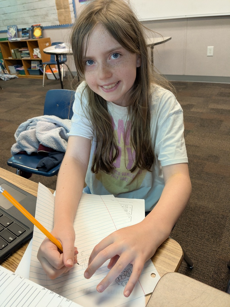 A student sits at a desk writing in a notebook with a pencil, looking up and smiling.