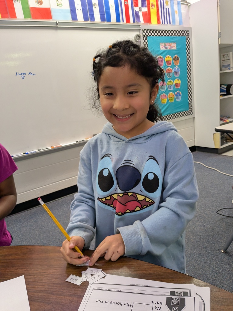 A student in a blue hoodie smiles while holding a pencil and working with word cards on a desk.