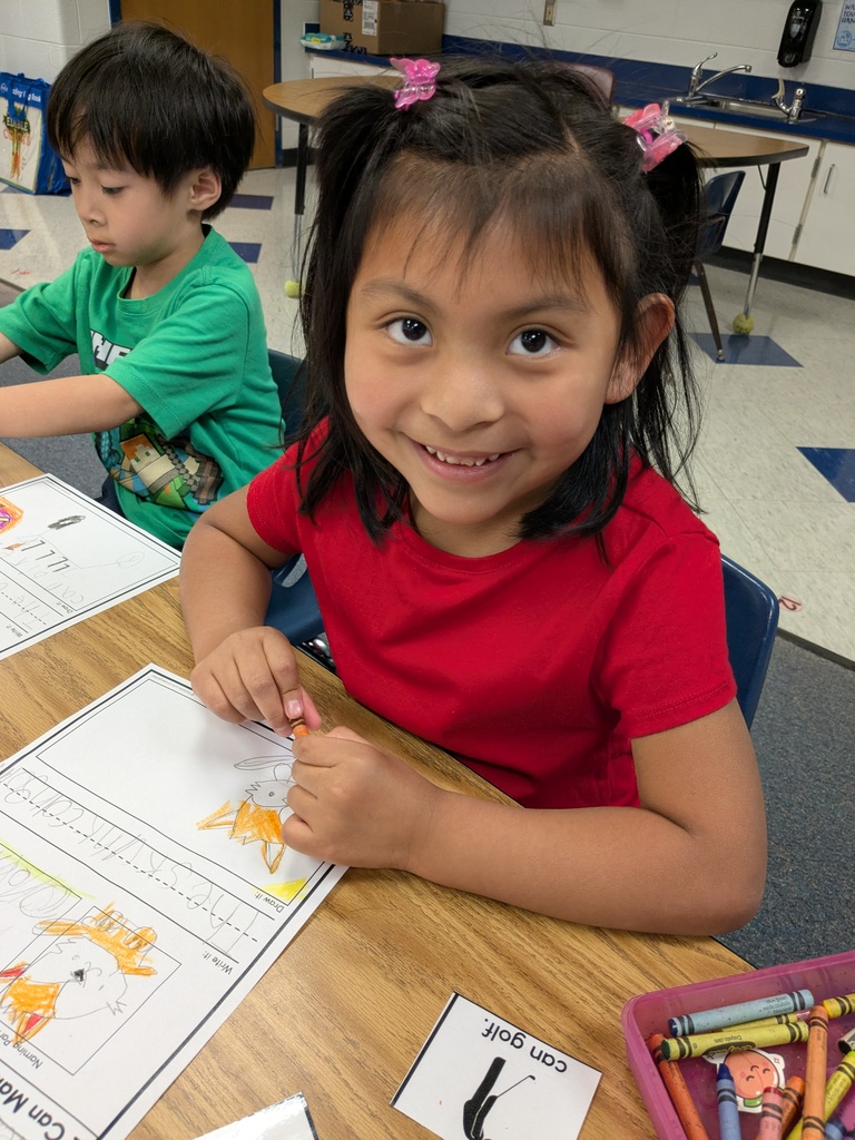A smiling student in a red shirt colors a worksheet at their desk, looking up toward the camera.