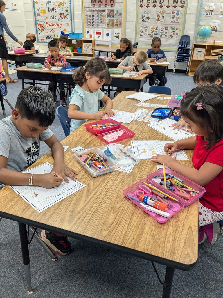 A group of elementary students sit at tables coloring and completing worksheets together during a class activity.