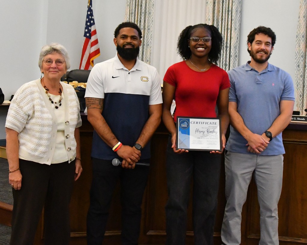 student holding certificate in board room between coaches with board member.  there is a US flag behind them 