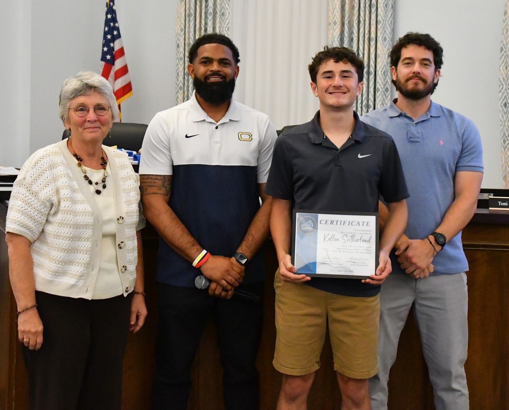 student holding certificate in board room between coaches with board member.  there is a US flag behind them 