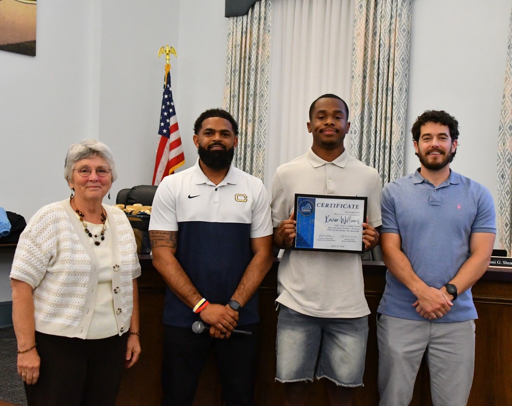 student holding certificate in board room between coaches with board member.  there is a US flag behind them 