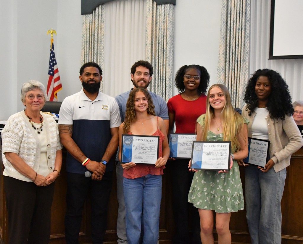 four students holding certificate in board room between coaches with board member.  there is a US flag behind them 
