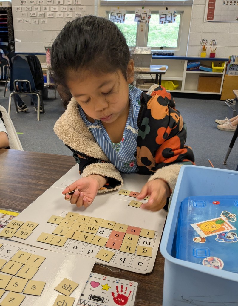 A young student sits at a classroom table using letter tiles to build words during a phonics activity, with other children working in the background.