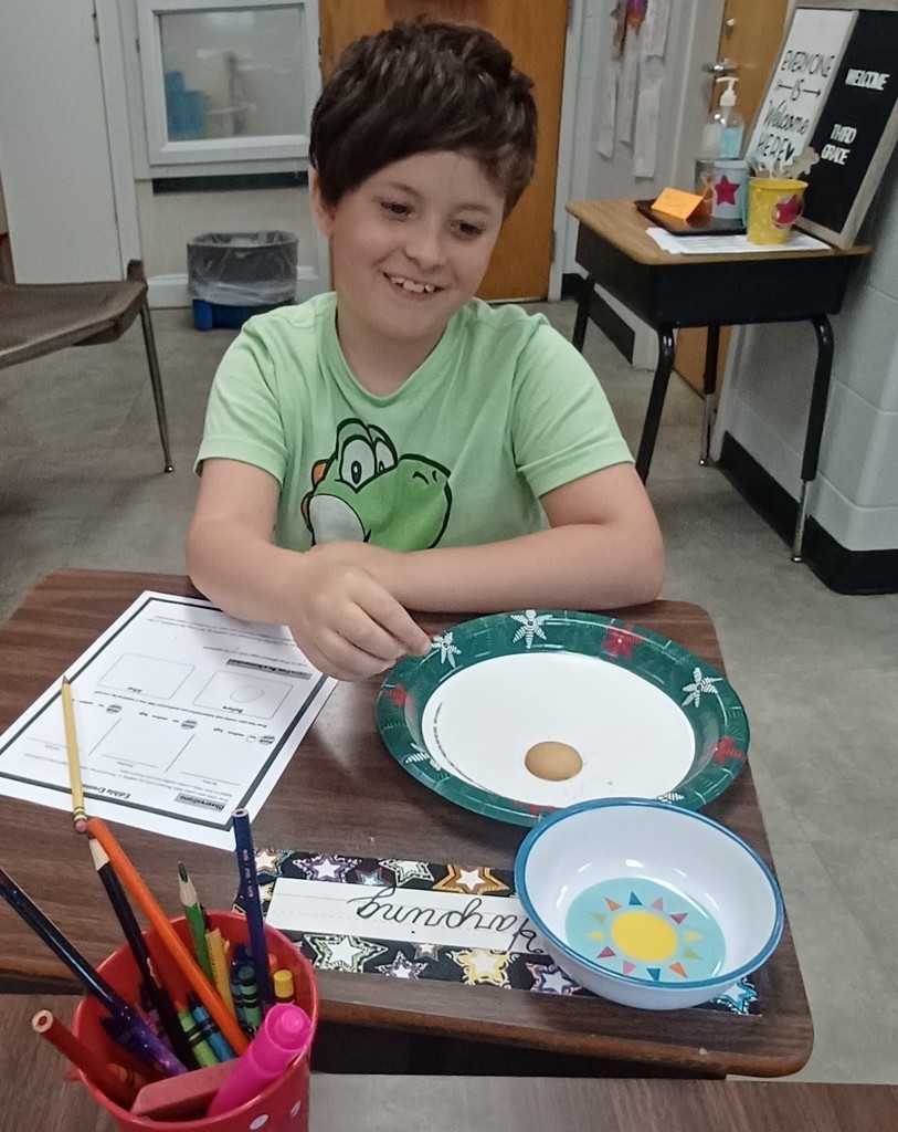 A smiling student in a green shirt sits at a desk, holding a small object over a paper plate with a cookie. A worksheet, bowl, and cup of pencils are on the desk, indicating a classroom experiment or observation activity.