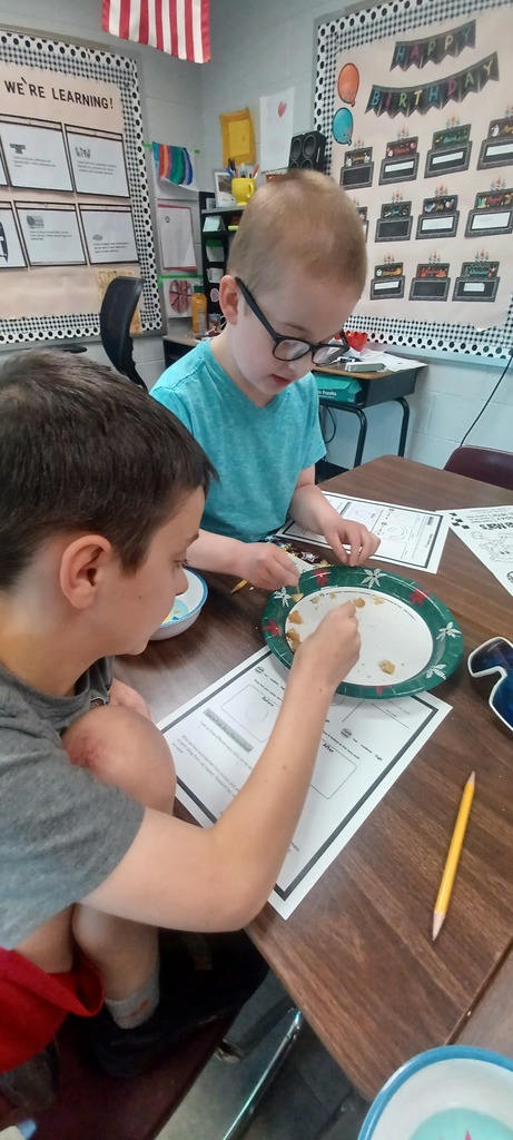 Two students sit side by side at a desk, using toothpicks to examine pieces of a cookie on a paper plate. A worksheet and pencil are in front of them as they work together on a classroom activity.