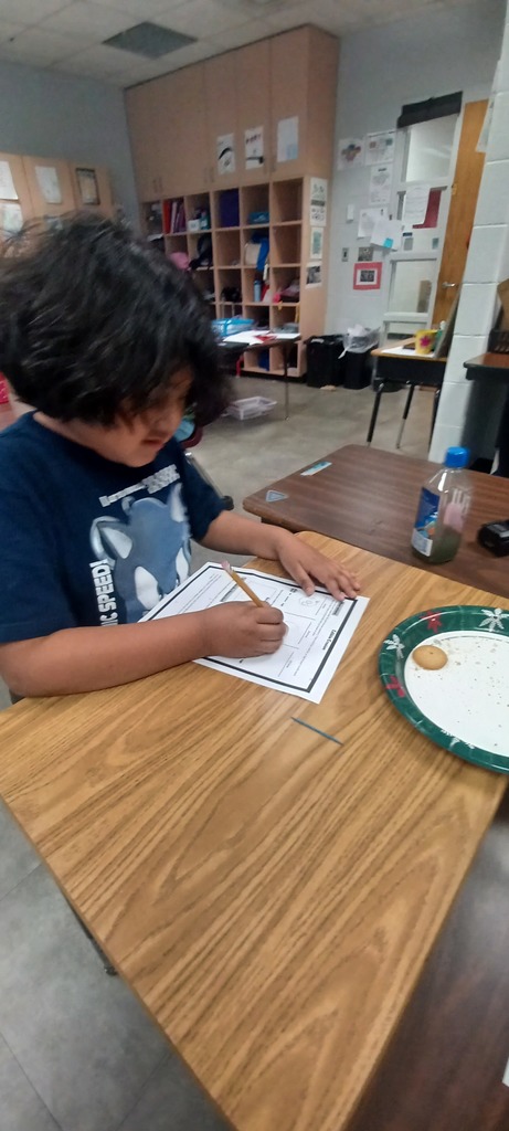 A student with long dark hair sits at a classroom desk, writing on a worksheet with a pencil. A paper plate with a cookie and crumbs sits nearby, along with a small bottle, suggesting a hands-on classroom activity.