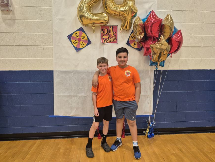 Two boys in orange shirts stand side by side smiling in front of a “24 Game” backdrop with balloons.