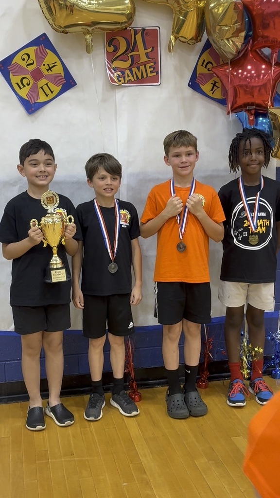 Four elementary-aged boys stand in a gym holding medals, one holding a trophy, in front of a “24 Game” backdrop with balloons.