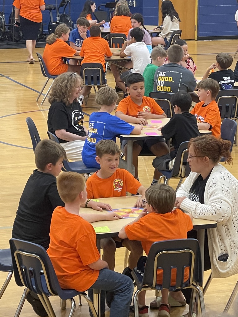 Groups of students sit at tables in a school gym playing a math card game, with teachers and volunteers assisting.