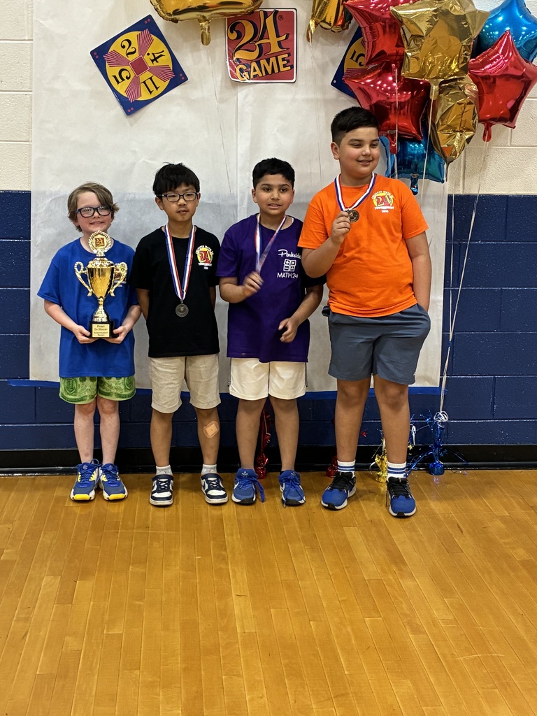 Four boys wearing medals pose in a gym; one holds a trophy while others stand beside him under “24 Game” decorations.