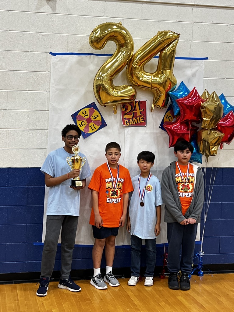 Four students pose in front of a backdrop with gold “24” balloons and colorful star balloons. One student holds a trophy, and the others wear medals, showing their achievements in the Math 24 competition.