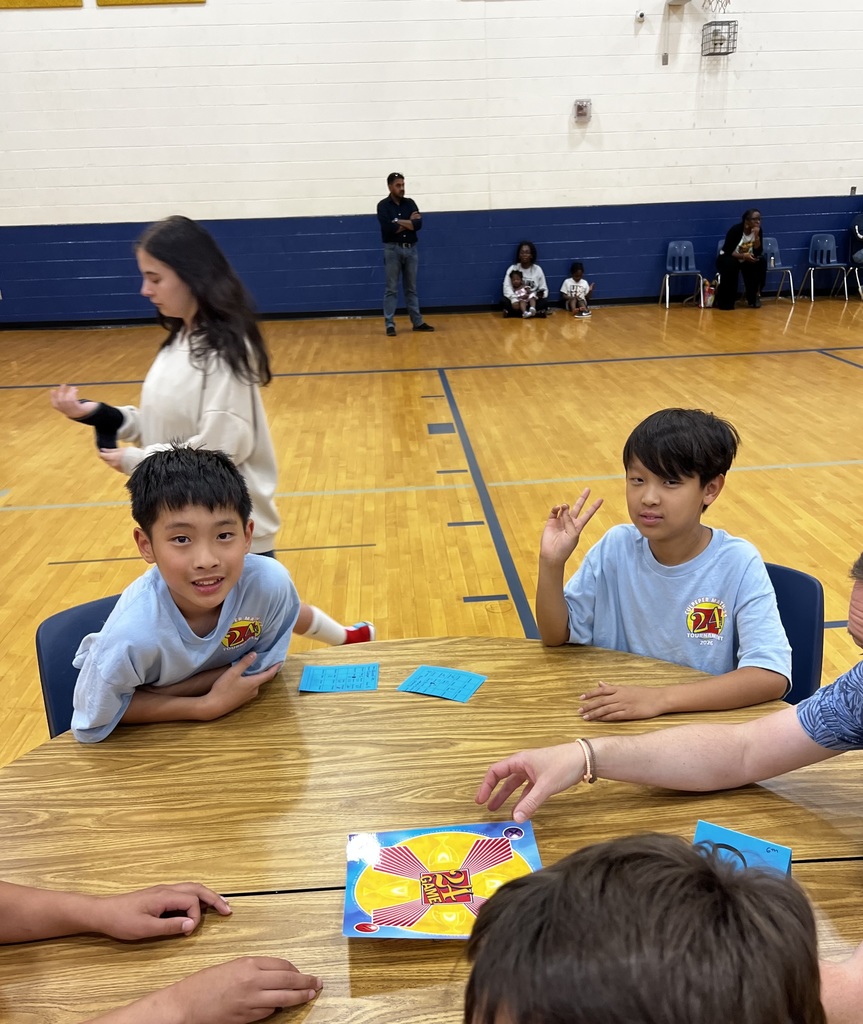 Two students sit at a table during a Math 24 game, with cards and the game board in front of them. One student smiles at the camera while the other makes a peace sign, with other participants nearby.