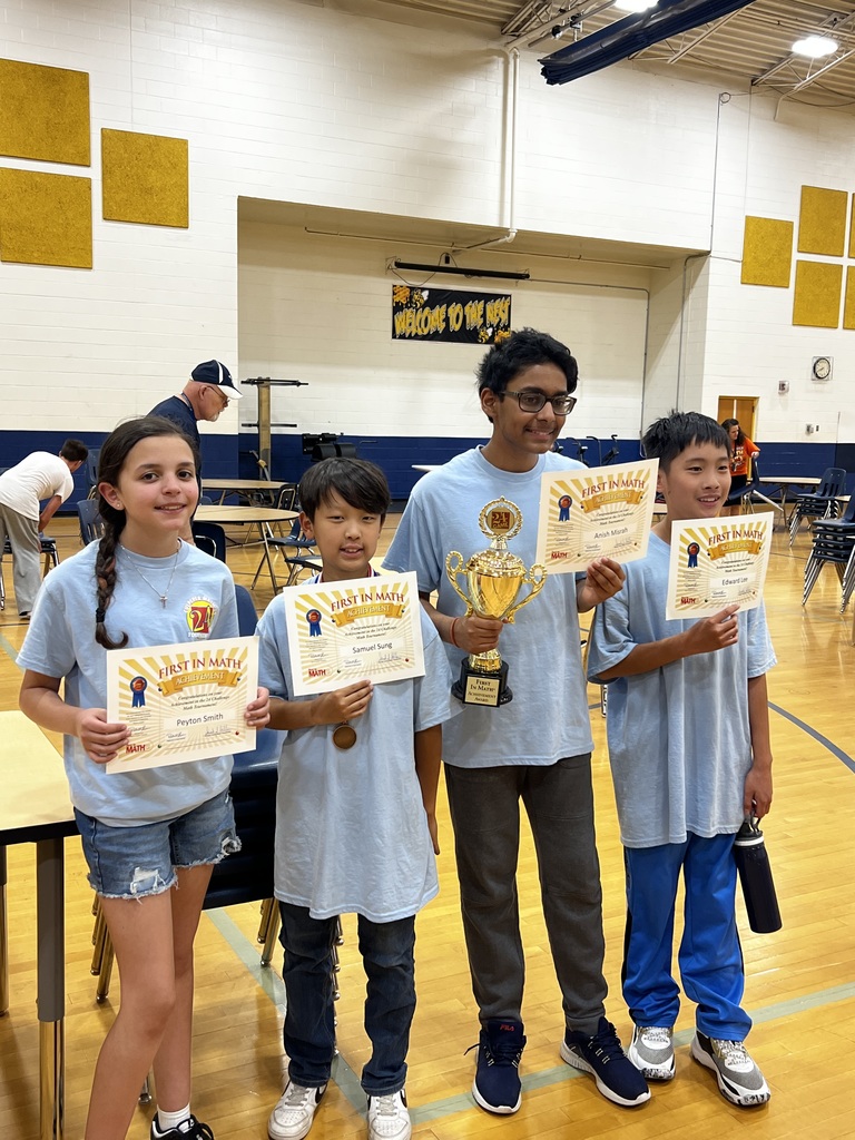 Four middle school students stand in a gym holding certificates from a Math 24 competition. One student holds a large trophy while all smile proudly. Tables and chairs are set up behind them.