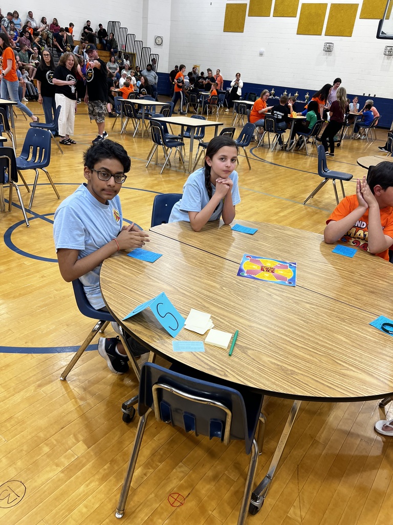 Students sit around tables in a school gym during a Math 24 competition. In the foreground, two students sit at a table with game cards, while many participants and spectators fill the background.