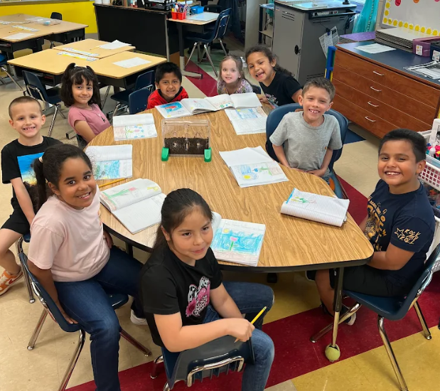 group of young students sit around a classroom table smiling, with open notebooks showing plant drawings and a small container in the center.