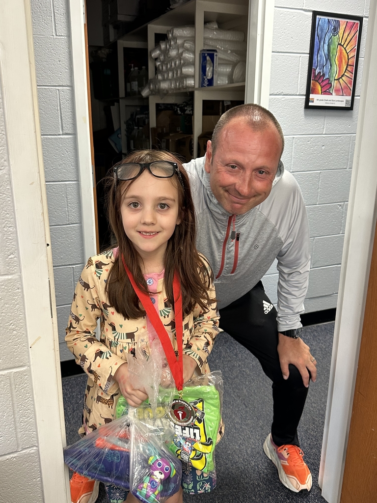  An indoor, eye-level shot captures a man and a young girl posing together in a doorway. The girl, with long brown hair and black-framed glasses resting on her head, stands on the left, smiling. She wears a tan long-sleeved shirt with a repeating print of dachshunds wearing colorful striped sweaters. A red ribbon with a circular medal hangs around her neck, and she holds a large plastic gift bag containing colorful toys and a neon green shirt.  Beside her, a man with short brown hair leans forward, resting his left hand on his knee. He wears a light gray quarter-zip jacket with red accents, black Adidas athletic pants, and bright orange sneakers. The background reveals a storage room filled with various supplies on shelves. A small, colorful piece of children's artwork featuring a sun and a flower is framed on the light blue cinderblock wall to the right.