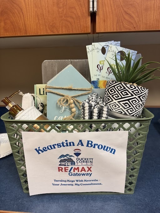    A green plastic basket filled with home‑themed gift items. Inside the basket are a small potted plant in a black‑and‑white geometric pot, a light blue wooden house‑shaped sign tied with twine, a rolled white towel, a gold soap dispenser, and a small decorative item with the word “Live.” Several brochures stand upright in the back. A label on the front reads: “Kearstin A Brown, Duckett & Corbin, RE/MAX Gateway — Turning Keys With Kearstin: Your Journey. My Commitment.”