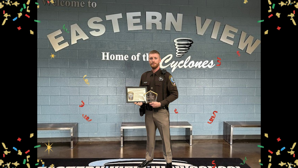 A school resource officer stands proudly in the lobby of Eastern View High School, centered in front of a large blue-gray brick wall that reads “Welcome to Eastern View, Home of the Cyclones.” He is wearing a dark brown sheriff’s uniform shirt with tan pants and black shoes. He holds a framed certificate in one hand and a clear, angular award plaque in the other, recognizing his achievement. Decorative confetti and streamers in gold, red, green, and blue are scattered around the border of the image, giving it a celebratory feel. Three metal benches line the wall behind him, and part of the school’s Cyclones logo is visible on the polished floor at his feet.