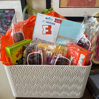 A white woven-pattern basket filled with assorted gift items sits on a table. Inside are orange gift bags, a box labeled “Fandango at Home” with a large “F,” packaged snacks, candy, and small treats wrapped in clear cellophane. Additional items, including a book and colorful packages, are tucked around the basket, creating a festive gift arrangement.
