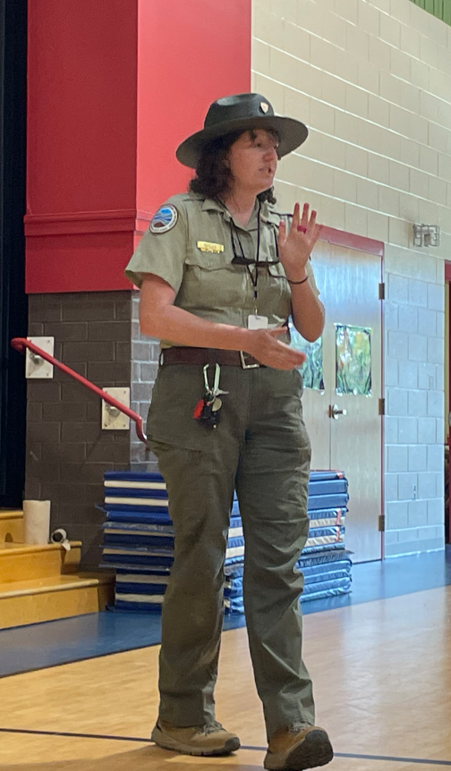 A park ranger stands in a gym speaking to students, gesturing while giving a presentation.