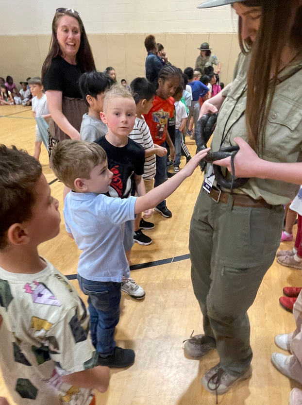 Children line up to take turns touching a snake held by a park ranger, with a teacher smiling nearby.