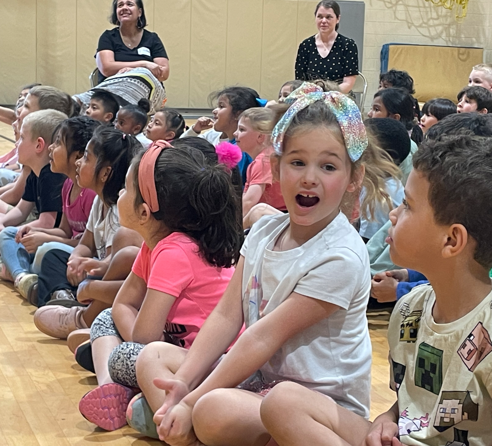 A group of children sit on the gym floor watching a presentation, with some looking excited and engaged.