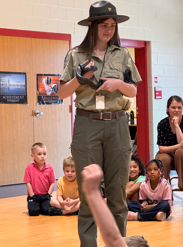 A park ranger stands in a school gym holding a snake while young children sit on the floor watching attentively.