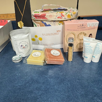 A collection of spa and self-care items is arranged on a table. The display includes a floral-patterned zippered bag, a black gift bag with tissue paper, and various skincare products. In front are wrapped bars of soap, a small jar (likely lip balm or salve), a pouch of bath soak, a reed diffuser with sticks, and two tubes of lotion or cream. The items are neatly organized, suggesting a gift set or relaxation-themed package.
