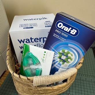 A woven basket contains dental care products arranged as a gift set. Inside is a boxed Waterpik water flosser, an Oral-B Pro 1000 rechargeable electric toothbrush, and a green mouth guard in its packaging. The items are neatly placed together, suggesting a practical oral hygiene-themed gift basket.