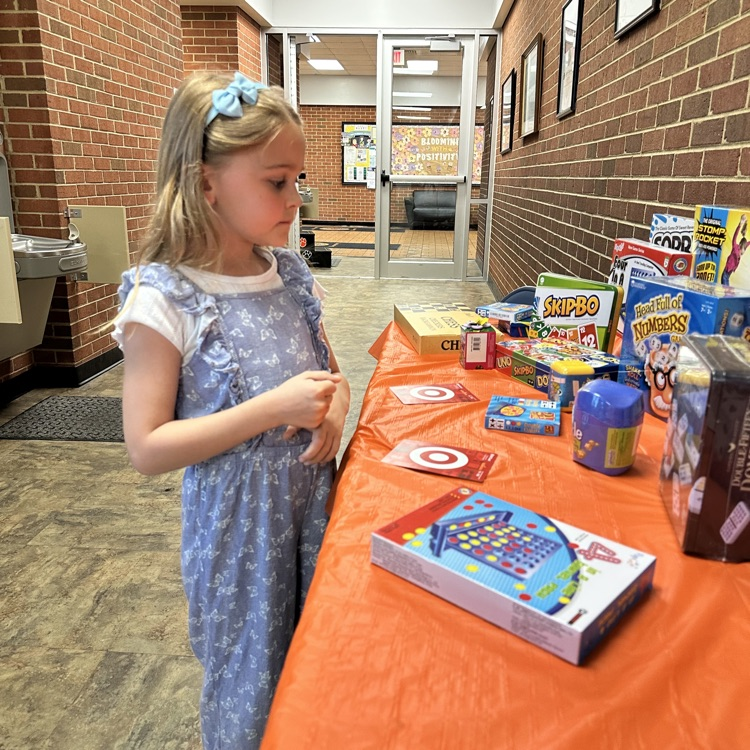     A young girl stands at a prize table covered in games and toys, looking thoughtfully at the selection.