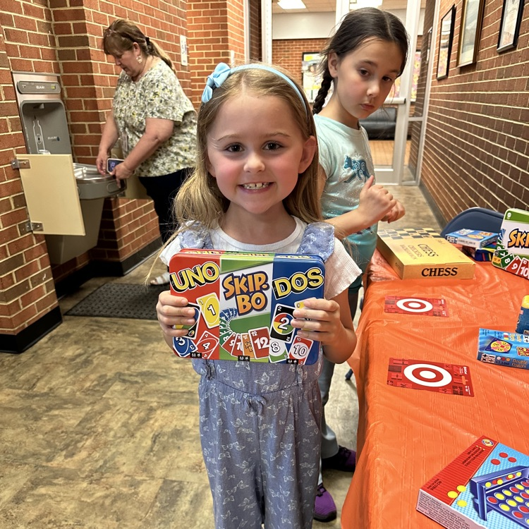     A smiling young girl holds up a prize box featuring UNO, Skip-Bo, and DOS card games while standing near the prize table.