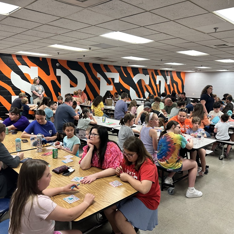     A wide view of a crowded school cafeteria filled with families and students playing bingo together at long tables.