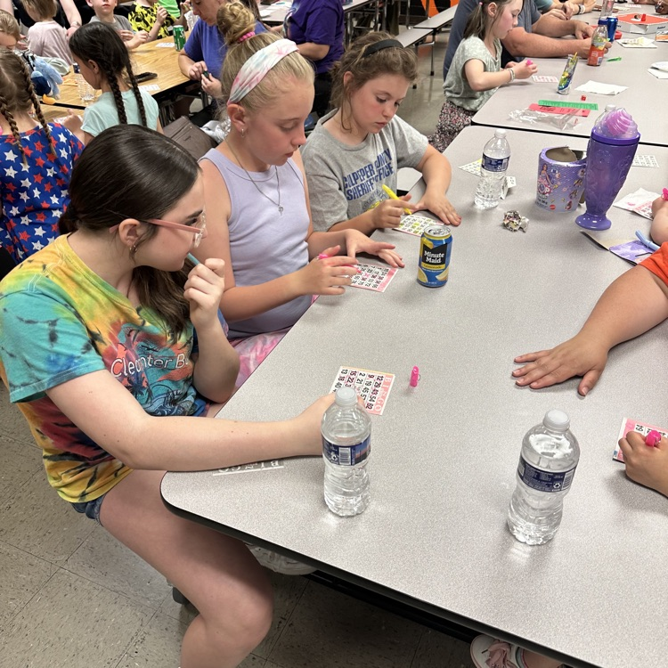     Several elementary-aged students sit side by side at a table, concentrating as they mark their bingo cards with bright markers.