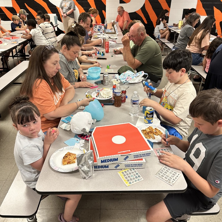     A group of students and adults sit together at cafeteria tables playing bingo, with pizza boxes and drinks on the table, focused on their cards.