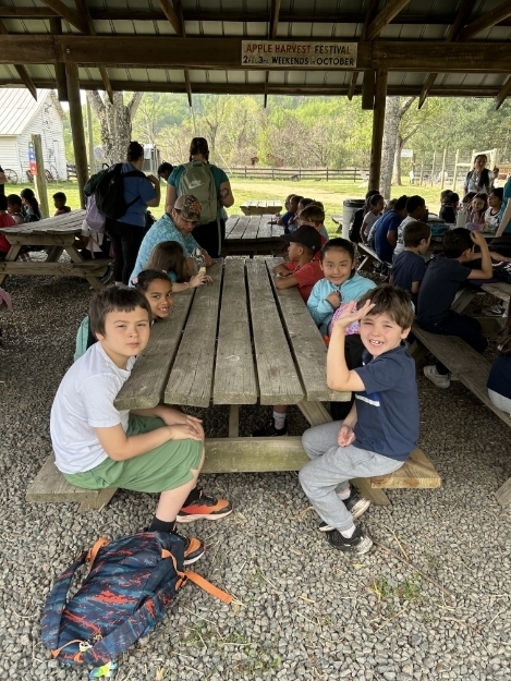 A group of elementary students sit at a wooden picnic table under a covered outdoor pavilion, talking and eating lunch. Backpacks and other students are visible in the background.